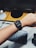 A person wearing a smartwatch on their wrist, with a digital display showing the time and various widgets. In the background, there is a wooden desk with a computer monitor, a round speaker, and a keyboard partially visible.