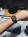 A person wearing a smartwatch on their wrist, with a digital display showing the time and various widgets. In the background, there is a wooden desk with a computer monitor, a round speaker, and a keyboard partially visible.