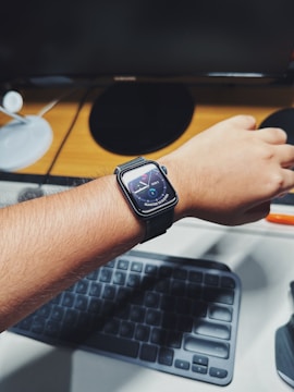 A person wearing a smartwatch with a colorful digital face showing the time. The wrist is positioned over a computer keyboard, with other items like a lamp and monitor in the background on a wooden desk.