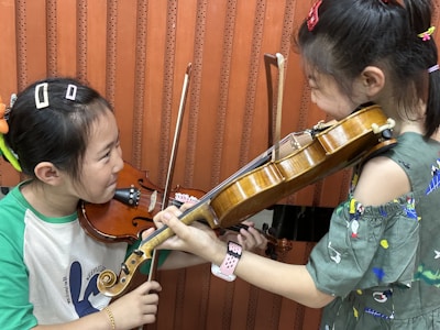Two children are playing violins facing each other. They both have bows in their hands, and they appear to be engaged in a playful or musical activity. The background consists of a wooden, orange-colored wall panel.