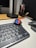 A digital watch with a colorful display is placed on top of a computer keyboard. The watch shows the time in large, bold, red and blue digital numbers. To the right, there are several pens standing in a container, and part of a computer monitor is visible in the background.