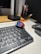 A digital watch with a colorful display is placed on top of a computer keyboard. The watch shows the time in large, bold, red and blue digital numbers. To the right, there are several pens standing in a container, and part of a computer monitor is visible in the background.