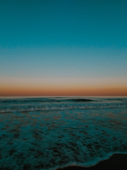 A tranquil ocean scene with gentle waves lapping against the shore under a vibrant sky. The sky transitions from a deep turquoise at the top to a warm orange near the horizon, suggesting a sunset or sunrise.