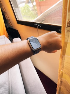 A person's wrist wearing a smartwatch is visible in a cozy room setting. The soft light coming through the window illuminates the area, highlighting the fabric strap of the watch. The room appears comfortable with curtains that gently filter the natural light.