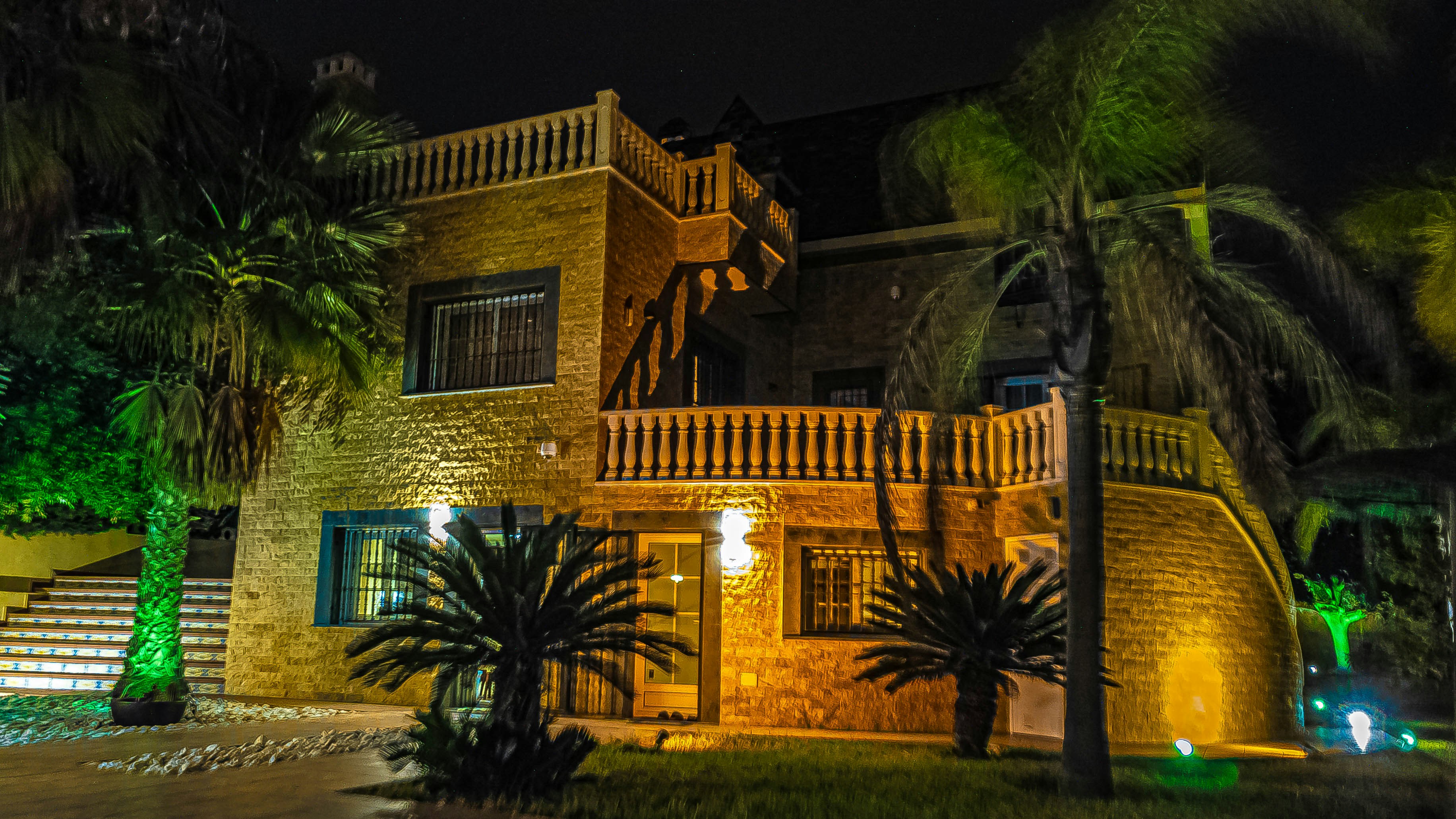 A large, luxurious house with warm lighting, surrounded by palm trees and greenery. The facade is constructed with stone and features ornate railing and balustrades. The scene is set at night, with strategic lighting illuminating parts of the building and vegetation, creating an inviting atmosphere.