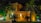 A large, luxurious house with warm lighting, surrounded by palm trees and greenery. The facade is constructed with stone and features ornate railing and balustrades. The scene is set at night, with strategic lighting illuminating parts of the building and vegetation, creating an inviting atmosphere.