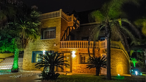 A large, luxurious house with warm lighting, surrounded by palm trees and greenery. The facade is constructed with stone and features ornate railing and balustrades. The scene is set at night, with strategic lighting illuminating parts of the building and vegetation, creating an inviting atmosphere.
