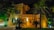 A large, luxurious house with warm lighting, surrounded by palm trees and greenery. The facade is constructed with stone and features ornate railing and balustrades. The scene is set at night, with strategic lighting illuminating parts of the building and vegetation, creating an inviting atmosphere.