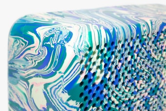 A close-up image of a textured surface that features vibrant, swirling patterns in shades of blue, green, and pink. The surface appears to have small, evenly spaced holes that add a three-dimensional effect. The design is reminiscent of marbled artwork, with smooth flowing lines and curves.