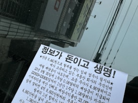 A close-up view of a piece of paper with Korean text, placed against a dark, reflective surface possibly a window. The background shows reflections of building structures and power lines.