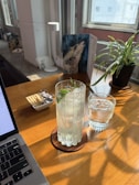 A sunlit workspace features a wooden table with a laptop partially in view. On the table, there is a tall glass filled with an iced beverage garnished with a sprig of rosemary, next to a smaller glass of water, both set on coasters. A small dish contains crackers and wrapped chocolates. A potted plant and a piece of art are visible near the window, enhancing the cozy atmosphere.