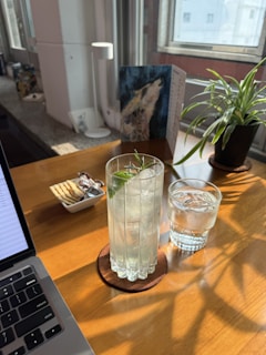 A sunlit workspace features a wooden table with a laptop partially in view. On the table, there is a tall glass filled with an iced beverage garnished with a sprig of rosemary, next to a smaller glass of water, both set on coasters. A small dish contains crackers and wrapped chocolates. A potted plant and a piece of art are visible near the window, enhancing the cozy atmosphere.