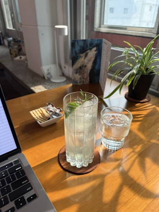A sunlit workspace features a wooden table with a laptop partially in view. On the table, there is a tall glass filled with an iced beverage garnished with a sprig of rosemary, next to a smaller glass of water, both set on coasters. A small dish contains crackers and wrapped chocolates. A potted plant and a piece of art are visible near the window, enhancing the cozy atmosphere.