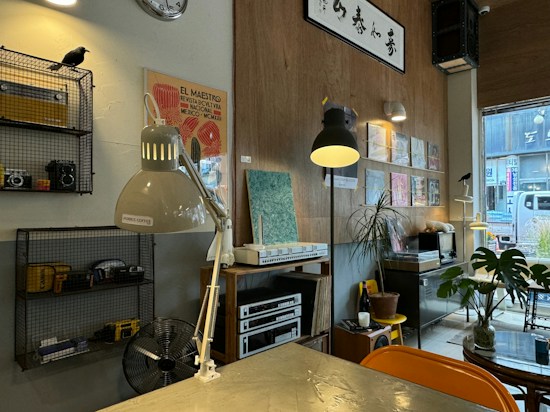 A cozy interior space featuring a mix of modern and vintage elements. On the left, a metal shelf holds cameras and a black crow ornament. Two desk lamps, one labeled 'MORRIS COFFEE,' and another provide lighting. A wall showcases an art piece with the text 'EL MAESTRO' and several mounted records and paintings. Below, a potted plant sits on a table amidst a variety of decor items. A window on the right shows a glimpse of the exterior street.