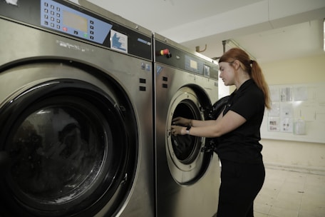 A person stands next to a commercial washing machine, inserting or removing laundry. The setting appears to be a laundry facility with tiled floors and walls. The washing machines are metallic with digital controls.