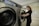 A person stands next to a commercial washing machine, inserting or removing laundry. The setting appears to be a laundry facility with tiled floors and walls. The washing machines are metallic with digital controls.