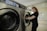 A person stands next to a commercial washing machine, inserting or removing laundry. The setting appears to be a laundry facility with tiled floors and walls. The washing machines are metallic with digital controls.