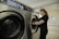 A person stands next to a commercial washing machine, inserting or removing laundry. The setting appears to be a laundry facility with tiled floors and walls. The washing machines are metallic with digital controls.