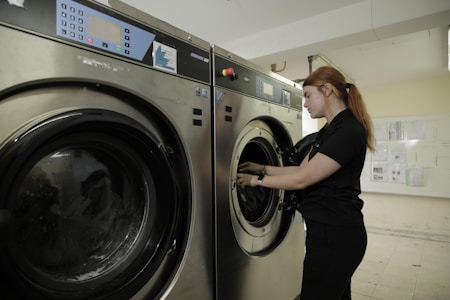 A person stands next to a commercial washing machine, inserting or removing laundry. The setting appears to be a laundry facility with tiled floors and walls. The washing machines are metallic with digital controls.