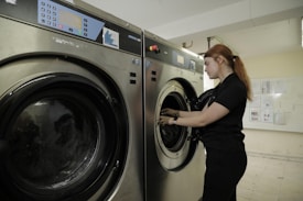 A person stands next to a commercial washing machine, inserting or removing laundry. The setting appears to be a laundry facility with tiled floors and walls. The washing machines are metallic with digital controls.