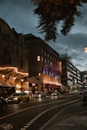 A street scene at dusk with illuminated buildings showcasing theaters or entertainment venues. Neon lights and signage glow warmly, highlighting architectural details. Vehicles, including a taxi, are seen on the road and a bike lane is marked in the foreground. A few people are present on the sidewalk.