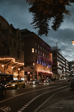 A street scene at dusk with illuminated buildings showcasing theaters or entertainment venues. Neon lights and signage glow warmly, highlighting architectural details. Vehicles, including a taxi, are seen on the road and a bike lane is marked in the foreground. A few people are present on the sidewalk.