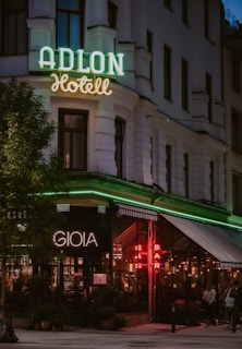 A corner building with a classic architectural style is prominently featured, showcasing a neon sign reading 'ADLON Hotell' with green lighting. Below, another sign spells 'GIOIA', beside a brightly lit 'BAR' sign in red. The setting is likely a hotel and bar, with glass windows revealing patrons gathered inside under warm, ambient lighting. The street scene is complemented by potted plants and softly lit exterior.