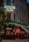 A corner building with a classic architectural style is prominently featured, showcasing a neon sign reading 'ADLON Hotell' with green lighting. Below, another sign spells 'GIOIA', beside a brightly lit 'BAR' sign in red. The setting is likely a hotel and bar, with glass windows revealing patrons gathered inside under warm, ambient lighting. The street scene is complemented by potted plants and softly lit exterior.