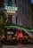 A corner building with a classic architectural style is prominently featured, showcasing a neon sign reading 'ADLON Hotell' with green lighting. Below, another sign spells 'GIOIA', beside a brightly lit 'BAR' sign in red. The setting is likely a hotel and bar, with glass windows revealing patrons gathered inside under warm, ambient lighting. The street scene is complemented by potted plants and softly lit exterior.
