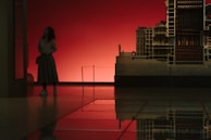 A person stands in a dimly lit room, looking at a large architectural model against a deep red background. The image is reflected on a polished floor, creating a symmetrical effect.