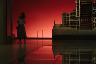 A person stands in a dimly lit room, looking at a large architectural model against a deep red background. The image is reflected on a polished floor, creating a symmetrical effect.