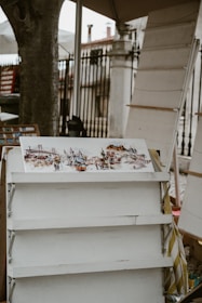 A watercolor painting of an urban scene is displayed on an easel. The artwork features a tram and some buildings, suggesting a street view from a city. The background includes some trees and architectural structures, possibly columns or rails, hinting at an outdoor location.