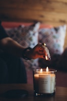 A hand with rings is lighting a scented candle labeled 'Cuir Nuit' in a cozy setting, with blurred colorful patterned cushions in the background and warm wooden tones.
