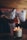 A hand with rings is lighting a scented candle labeled 'Cuir Nuit' in a cozy setting, with blurred colorful patterned cushions in the background and warm wooden tones.