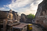 A serene stone statue of Buddha sits in a meditative pose on a raised platform amidst ancient ruins. The surrounding walls are partially intact with visible brickwork. The sky is clear with some wispy clouds, and there's a yellow donation box in the foreground.