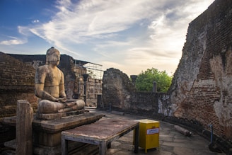 A serene stone statue of Buddha sits in a meditative pose on a raised platform amidst ancient ruins. The surrounding walls are partially intact with visible brickwork. The sky is clear with some wispy clouds, and there's a yellow donation box in the foreground.