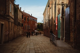 A narrow cobblestone street lined with historic brick buildings, some featuring colorful banners. A small group of people walks down the street, with the sun casting warm light and shadows. The scene appears quiet and serene, capturing an old-world charm.