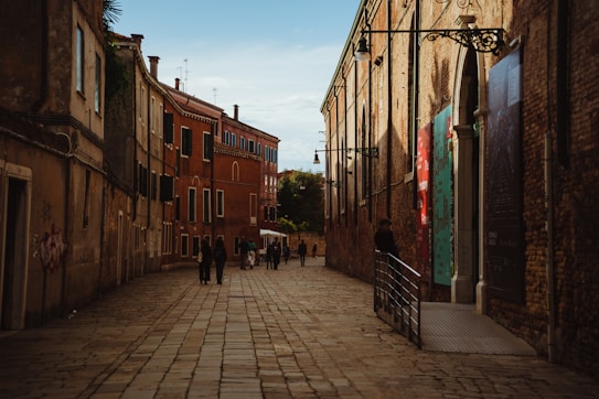 A narrow cobblestone street lined with historic brick buildings, some featuring colorful banners. A small group of people walks down the street, with the sun casting warm light and shadows. The scene appears quiet and serene, capturing an old-world charm.