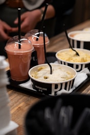 A wooden table holds trays with containers of food and drinks. The foreground features cups of pasta topped with cheese, accompanied by straws. Behind them are drinks with pinkish smoothies. The setting appears casual, suggesting a food stall.