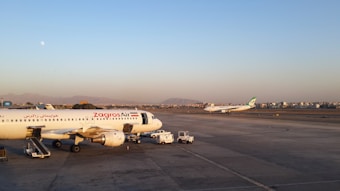 Airplanes are stationed on an airport tarmac, with one aircraft marked 'Zagros Air' prominently in the foreground. The background features another plane preparing for takeoff. The scene is set against a backdrop of distant mountains and a cityscape under a clear sky.