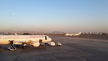 Airplanes are stationed on an airport tarmac, with one aircraft marked 'Zagros Air' prominently in the foreground. The background features another plane preparing for takeoff. The scene is set against a backdrop of distant mountains and a cityscape under a clear sky.