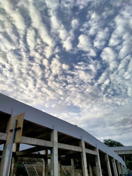 A cloudy sky with soft, dispersed white clouds is visible above a partially constructed or open-sided building with concrete columns and beams. The structure features a large rectangular opening, and in the background, a silhouette of trees is visible.