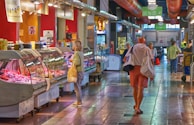 A bustling indoor market scene with people walking and browsing various food stalls. The setting features a long corridor with tiled flooring and overhead lighting. Stalls display a variety of meats inside glass cases. Several customers are engaging with stall vendors or walking down the aisle, carrying shopping bags.