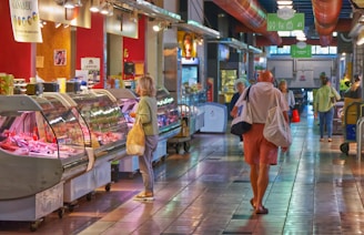 A bustling indoor market scene with people walking and browsing various food stalls. The setting features a long corridor with tiled flooring and overhead lighting. Stalls display a variety of meats inside glass cases. Several customers are engaging with stall vendors or walking down the aisle, carrying shopping bags.