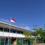 A school courtyard with a flagpole displaying a red and white flag. A person in a uniform stands near the flag. The school building in the background has a light blue roof and is surrounded by a green fence. Lush green trees and clear blue sky add to the scene.