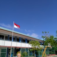 A school courtyard with a flagpole displaying a red and white flag. A person in a uniform stands near the flag. The school building in the background has a light blue roof and is surrounded by a green fence. Lush green trees and clear blue sky add to the scene.