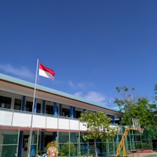 A school courtyard with a flagpole displaying a red and white flag. A person in a uniform stands near the flag. The school building in the background has a light blue roof and is surrounded by a green fence. Lush green trees and clear blue sky add to the scene.