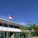 A school courtyard with a flagpole displaying a red and white flag. A person in a uniform stands near the flag. The school building in the background has a light blue roof and is surrounded by a green fence. Lush green trees and clear blue sky add to the scene.