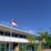 A school courtyard with a flagpole displaying a red and white flag. A person in a uniform stands near the flag. The school building in the background has a light blue roof and is surrounded by a green fence. Lush green trees and clear blue sky add to the scene.