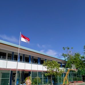 A school courtyard with a flagpole displaying a red and white flag. A person in a uniform stands near the flag. The school building in the background has a light blue roof and is surrounded by a green fence. Lush green trees and clear blue sky add to the scene.